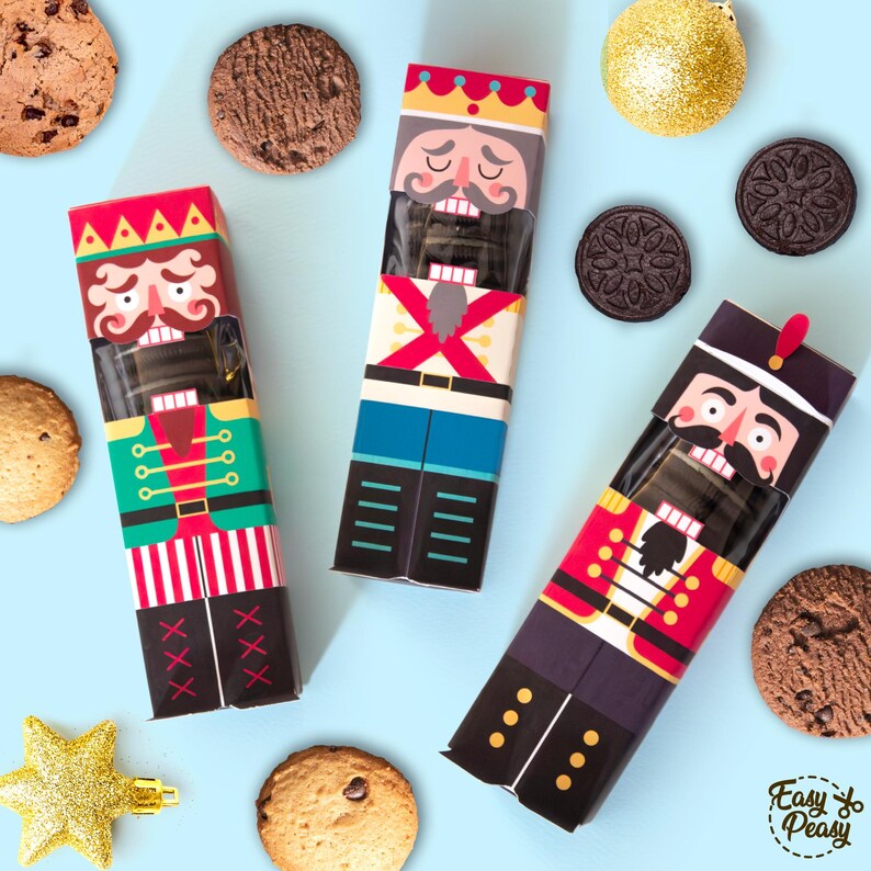 May include: Three nutcracker-themed cookie boxes with colorful designs, surrounded by chocolate chip cookies, chocolate sandwich cookies, and gold Christmas ornaments on a light blue background. The boxes feature red, green, and black uniforms.