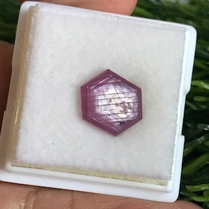 May include: A hexagonal, pink gemstone is presented in a white box. The gem is faceted and has a translucent quality, with internal features visible. The box is set against a blurred green backdrop.