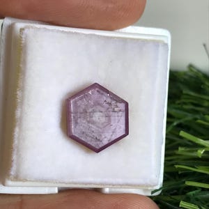 May include: A hexagonal, light purple gemstone with a flat, polished surface featuring a distinct internal geometric pattern. The stone is set within a square white display box with a soft, padded interior, showcasing its unique shape and colour.