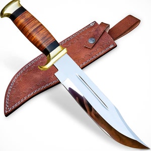 May include: A large, polished steel knife with a brown leather handle and a brass bolster. The knife is paired with a brown leather sheath with decorative stitching.