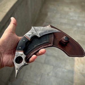 May include: A silver and black karambit knife with a brown leather sheath. The knife has a unique, curved blade and a finger ring.