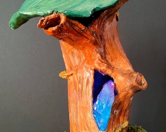 May include: A ceramic sculpture of a tree trunk with a green leafy top. The trunk has a hollow space with a blue crystal inside. The base of the sculpture is decorated with green moss, a red mushroom, and a small green frog.