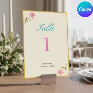 Puede incluir: Una tarjeta de número de mesa color crema con borde dorado, que muestra la palabra "Table" en escritura turquesa y el número "1" en rosa. También hay detalles florales y los nombres "Ethan & Lily" con la fecha "21 de septiembre de 2025". La tarjeta se sostiene en un soporte de metal plateado.
