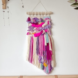 Può includere: Un arazzo da parete tessuto colorato con filato rosa, viola e bianco. L'arazzo ha un'asta di legno nella parte superiore ed è decorato con nappe.