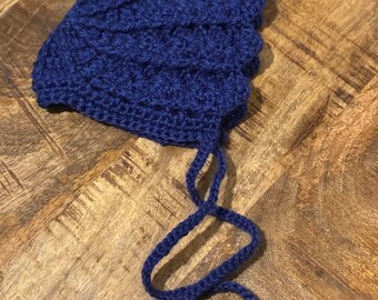 May include: A navy blue crochet bonnet with a textured stitch pattern. The bonnet has long ties for securing under the chin. The handmade hat is displayed on a wooden surface.