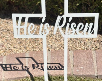 May include: A white, cut-out cross with the words "He is Risen" in a decorative font. The cross is placed in a grassy area with a brick border and a green bush in the background. The cross casts a shadow on the ground.