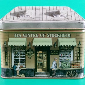 Może przedstawiać: Prostokątna puszka na herbatę z szczegółową ilustracją sklepu Tea Centre of Stockholm. Puszka ma konstrukcję dachu i przedstawia sklep z markizami, oknami i postaciami. Tekst "TEA CENTRE OF STOCKHOLM" jest wyświetlany z przodu.