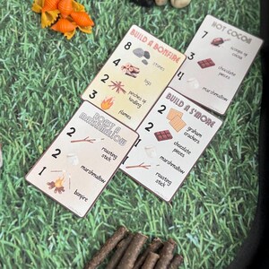 Puede incluir: Tarjetas con ilustraciones de camping, con instrucciones para construir una fogata, asar malvaviscos y preparar s'mores. Las tarjetas est&aacute;n sobre una superficie verde con hierba, con piedras, pasta y palitos.