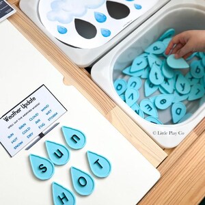 May include: A weather-themed educational activity set featuring a white board with a cloud illustration, a bin filled with light blue letter-shaped water droplets, and a card labeled Weather Update with words like Sun, Rain, and Cloud.