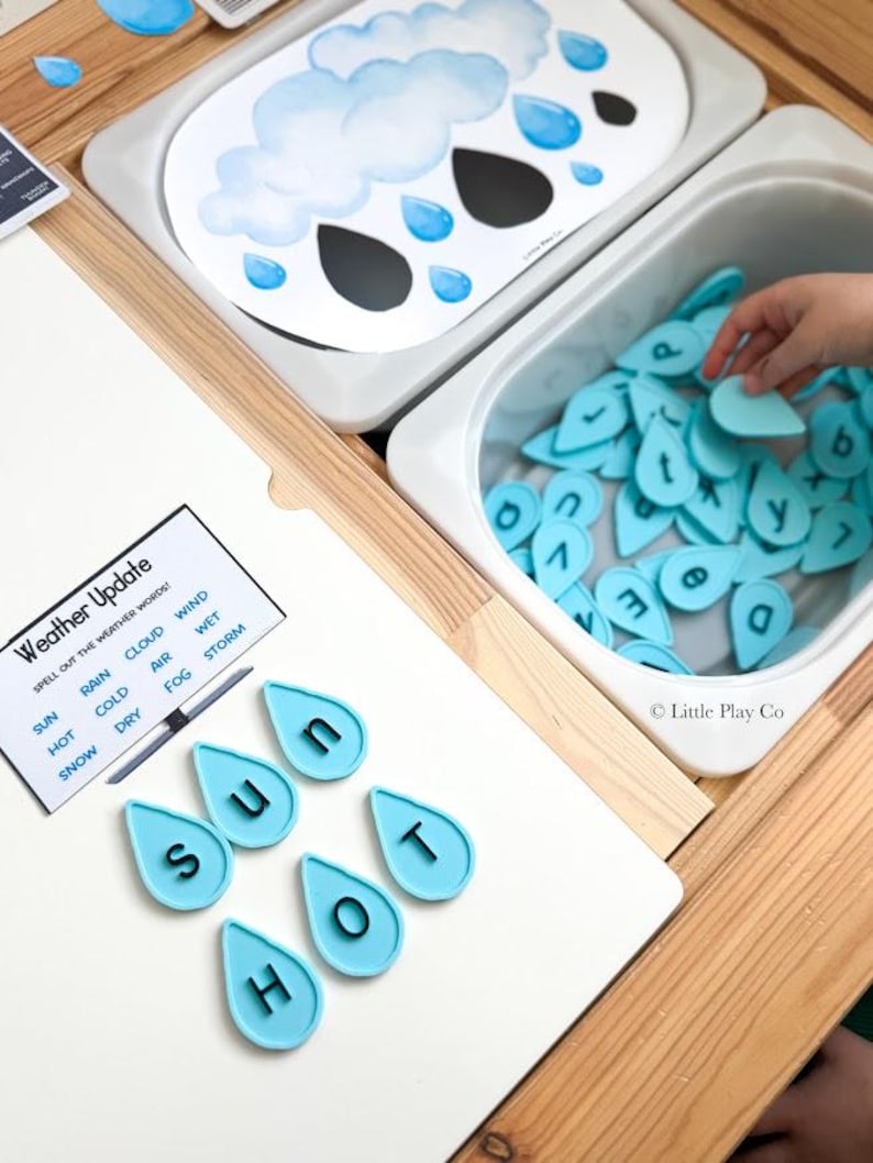 May include: A weather-themed educational activity set featuring a white board with a cloud illustration, a bin filled with light blue letter-shaped water droplets, and a card labeled Weather Update with words like Sun, Rain, and Cloud.