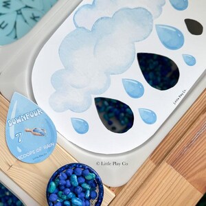 May include: A sensory play activity set featuring a cloud and raindrop-themed design. The set includes a white tray with a cloud and raindrop illustration, a blue scoop filled with blue beads, and a raindrop-shaped card with the text "DOWNPOUR".