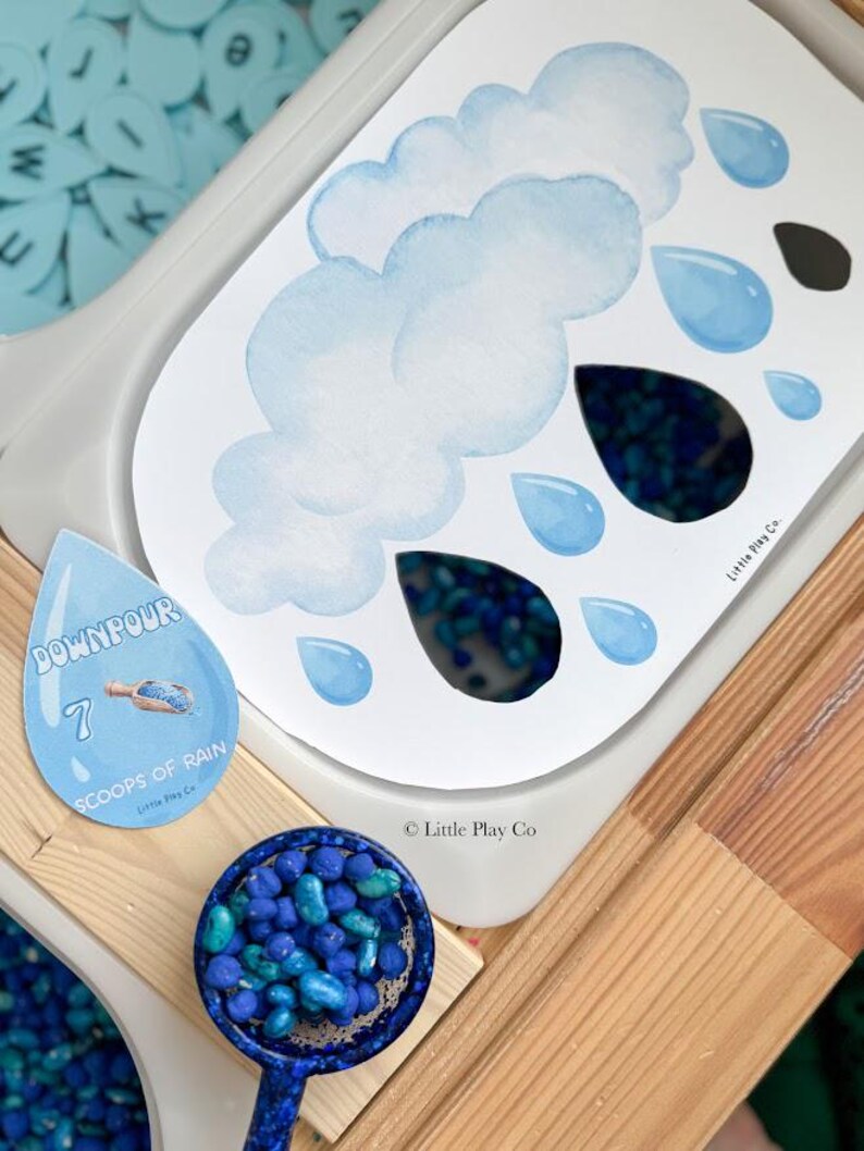 May include: A sensory play activity set featuring a cloud and raindrop-themed design. The set includes a white tray with a cloud and raindrop illustration, a blue scoop filled with blue beads, and a raindrop-shaped card with the text "DOWNPOUR".