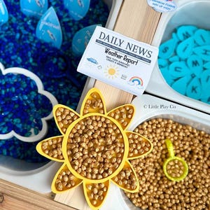 May include: A sensory play setup with a weather theme. Includes blue water beads, blue raindrop-shaped number cards, a yellow sun, and a newspaper-style "Weather Report" card. A rainbow and letter tiles are also present.