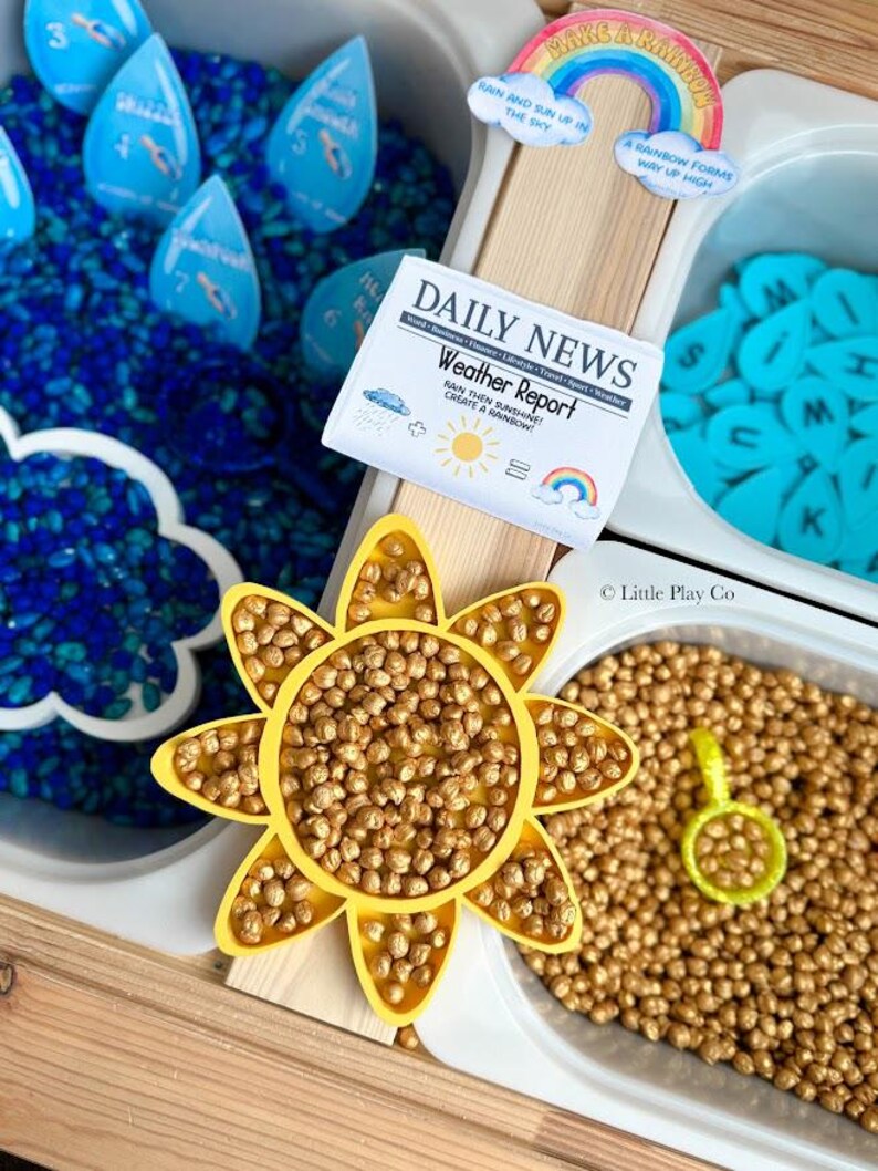 May include: A sensory play setup with a weather theme. Includes blue water beads, blue raindrop-shaped number cards, a yellow sun, and a newspaper-style "Weather Report" card. A rainbow and letter tiles are also present.