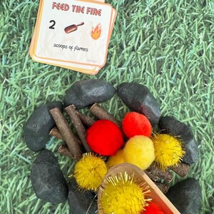 Puede incluir: Un juego infantil con tem&aacute;tica de fogata. El juego incluye una tarjeta que dice "Feed the Fire" con el n&uacute;mero 2, una cuchara de madera, pompones rojos y amarillos, palitos y piedras negras, todo dispuesto sobre una superficie verde.