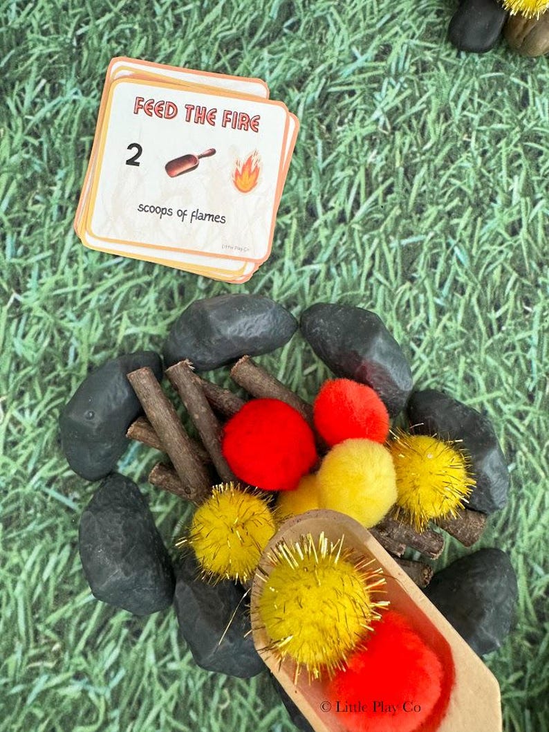 Puede incluir: Un juego infantil con tem&aacute;tica de fogata. El juego incluye una tarjeta que dice "Feed the Fire" con el n&uacute;mero 2, una cuchara de madera, pompones rojos y amarillos, palitos y piedras negras, todo dispuesto sobre una superficie verde.