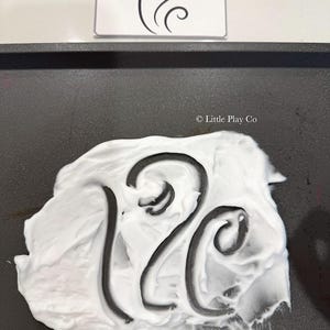 Puede incluir: Una bandeja negra con crema de afeitar blanca y el n&uacute;mero 12 negro dibujado en la crema. Una tarjeta blanca con un dise&ntilde;o abstracto negro est&aacute; encima de la bandeja.