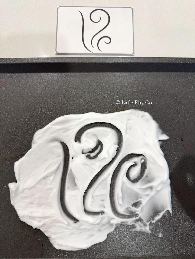 Puede incluir: Una bandeja negra con crema de afeitar blanca y el n&uacute;mero 12 negro dibujado en la crema. Una tarjeta blanca con un dise&ntilde;o abstracto negro est&aacute; encima de la bandeja.