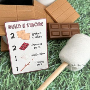 Puede incluir: Un conjunto de ingredientes de juguete para s'mores, que incluye galletas Graham, trozos de chocolate, una malvavisco en un palo y una tarjeta que dice "Build a S'more". Los art&iacute;culos est&aacute;n dispuestos sobre una superficie verde.
