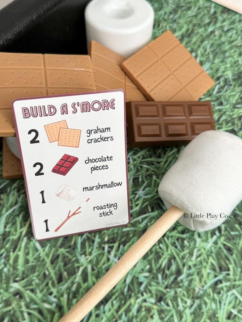 Puede incluir: Un conjunto de ingredientes de juguete para s'mores, que incluye galletas Graham, trozos de chocolate, una malvavisco en un palo y una tarjeta que dice "Build a S'more". Los art&iacute;culos est&aacute;n dispuestos sobre una superficie verde.