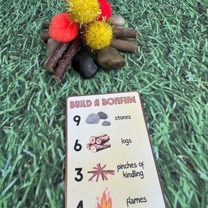 Puede incluir: Una escena de fogata en miniatura con pompones amarillos y rojos, palitos y piedras. Una tarjeta con el texto "BUILD A BONFIRE" muestra los componentes necesarios: piedras, troncos, yesca y llamas. El fondo es verde.