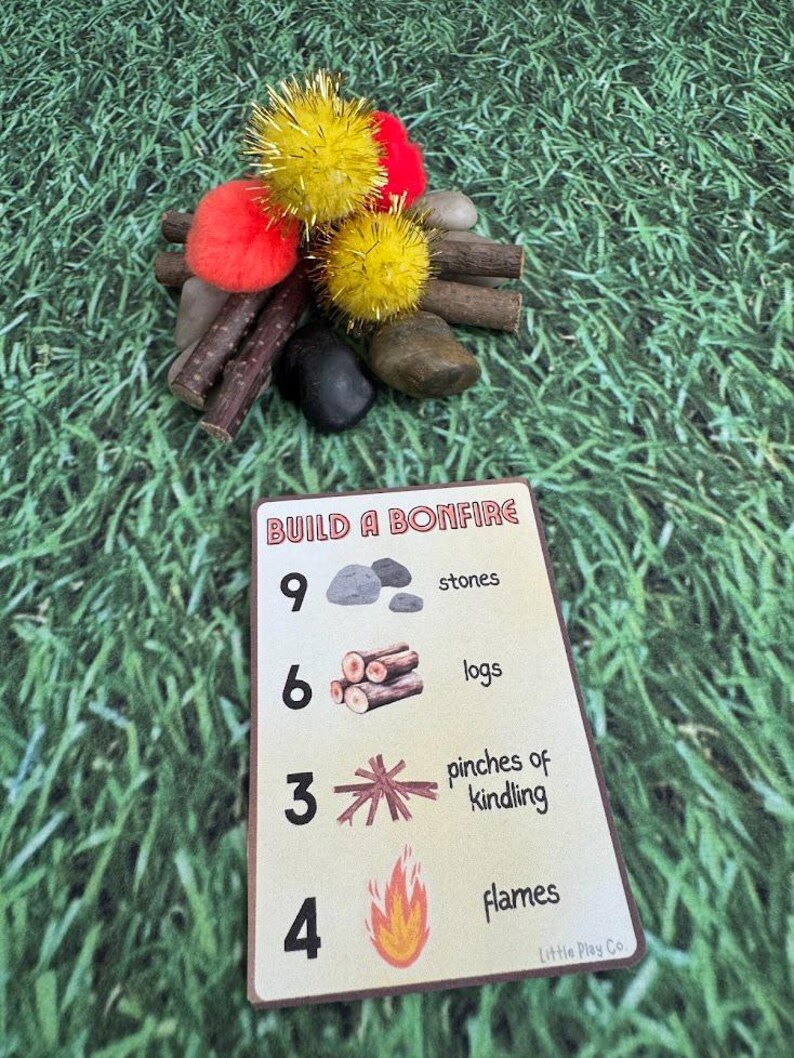 Puede incluir: Una escena de fogata en miniatura con pompones amarillos y rojos, palitos y piedras. Una tarjeta con el texto "BUILD A BONFIRE" muestra los componentes necesarios: piedras, troncos, yesca y llamas. El fondo es verde.