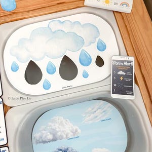 May include: Two oval educational activity mats featuring weather themes. The top mat displays watercolor clouds and raindrops with a Storm Alert card. The bottom mat illustrates various cloud types labeled Cirrus, Cumulus, Stratus, and Nimbus.