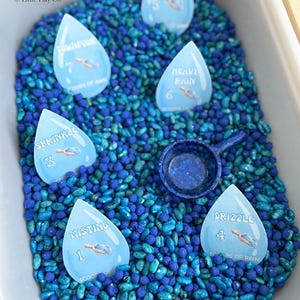May include: A sensory bin filled with blue and teal beans, featuring raindrop-shaped cards labeled with rain intensity terms like "Misting" and "Heavy Rain." A small blue scoop is also included, perfect for educational play.
