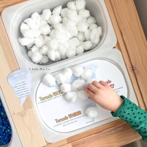 May include: A wooden sensory table with two white bins. One bin is filled with white cotton balls. The other bin contains a printed card labeled Tornado WATCH and Tornado WARNING with text explaining tornado safety and white cotton balls.