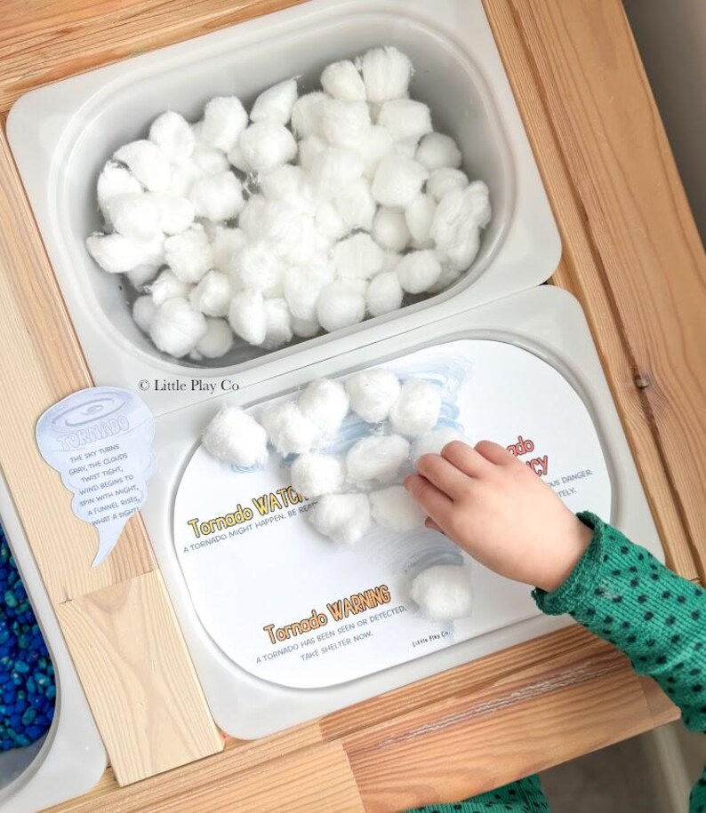 May include: A wooden sensory table with two white bins. One bin is filled with white cotton balls. The other bin contains a printed card labeled Tornado WATCH and Tornado WARNING with text explaining tornado safety and white cotton balls.