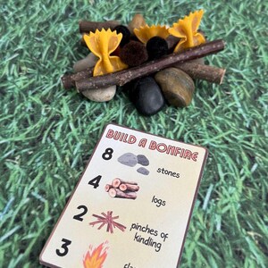 Puede incluir: Una escena de fogata en miniatura con llamas de pasta de lazo, ramitas, piedras y pompones. Tambi&eacute;n hay una tarjeta con las instrucciones "Build a Bonfire", con ilustraciones de piedras, troncos, yesca y llamas.