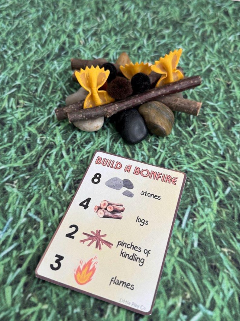 Puede incluir: Una escena de fogata en miniatura con llamas de pasta de lazo, ramitas, piedras y pompones. Tambi&eacute;n hay una tarjeta con las instrucciones "Build a Bonfire", con ilustraciones de piedras, troncos, yesca y llamas.