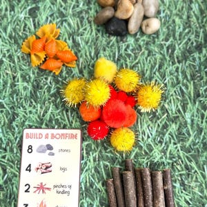 Puede incluir: Una colecci&oacute;n de materiales de manualidades para una actividad con tem&aacute;tica de hoguera. Incluye piedras peque&ntilde;as, pasta naranja, pompones amarillos y rojos, palitos de madera y una tarjeta con instrucciones. La tarjeta dice "Build a Bonfire".
