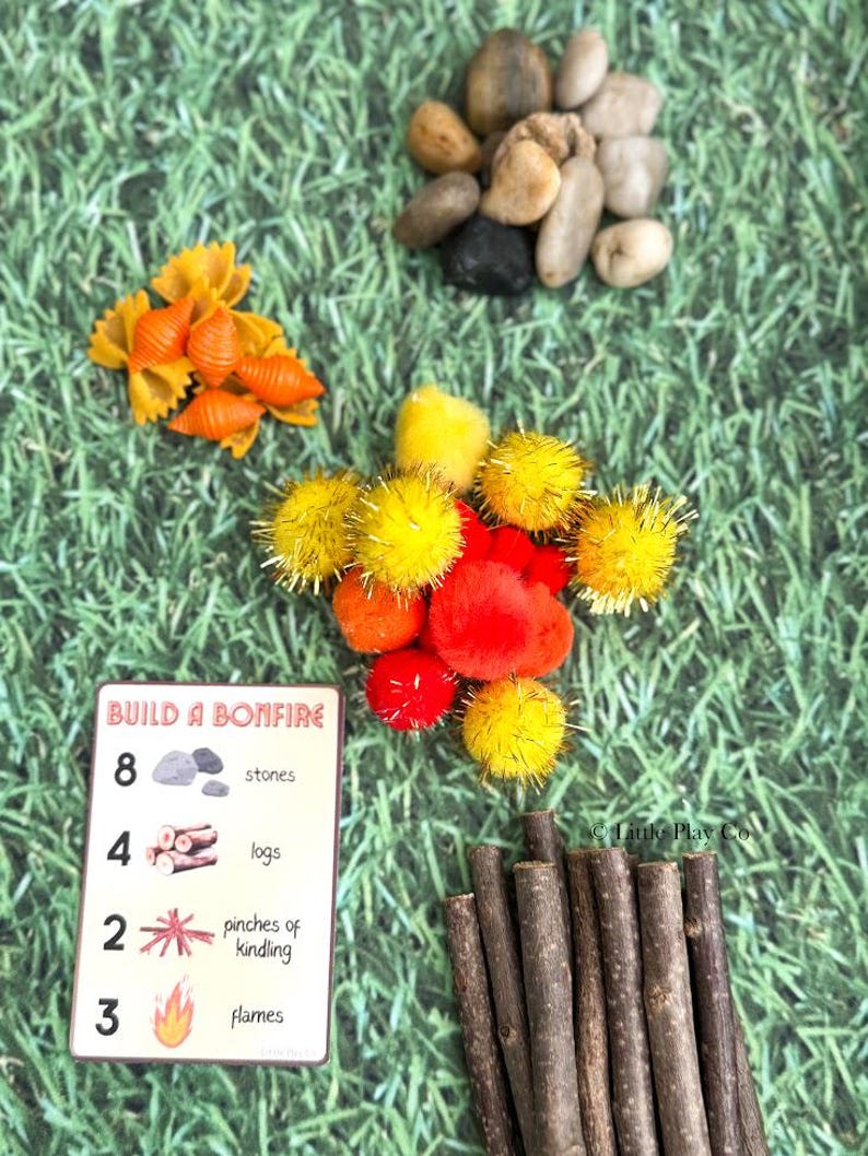 Puede incluir: Una colecci&oacute;n de materiales de manualidades para una actividad con tem&aacute;tica de hoguera. Incluye piedras peque&ntilde;as, pasta naranja, pompones amarillos y rojos, palitos de madera y una tarjeta con instrucciones. La tarjeta dice "Build a Bonfire".