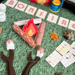 Puede incluir: Un juego de campamento de fieltro con la palabra "BONFIRE" escrita en baldosas de letras individuales. Incluye una fogata de fieltro rojo, una linterna azul, troncos de fieltro, malvaviscos en palitos y peque&ntilde;as rocas, todo dispuesto sobre una superficie verde.