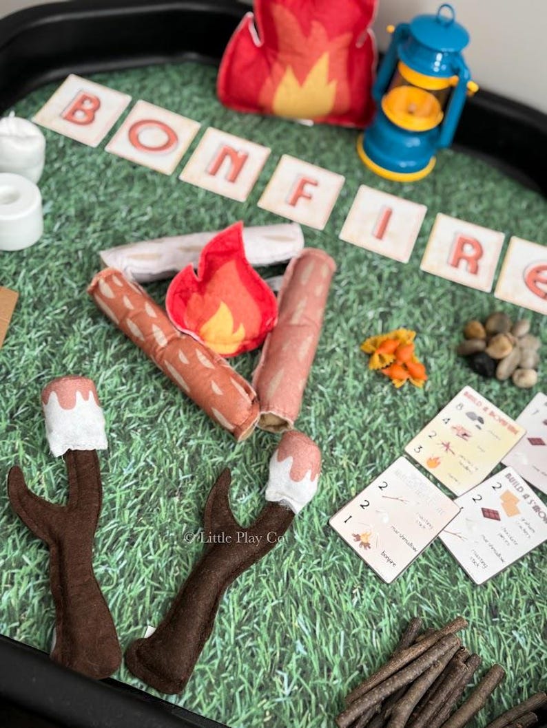 Puede incluir: Un juego de campamento de fieltro con la palabra "BONFIRE" escrita en baldosas de letras individuales. Incluye una fogata de fieltro rojo, una linterna azul, troncos de fieltro, malvaviscos en palitos y peque&ntilde;as rocas, todo dispuesto sobre una superficie verde.
