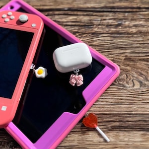 May include: A white wireless charging case with a pink bow charm attached. The case is sitting on top of a pink and black tablet case. A pink and white egg charm is attached to a pink and black Nintendo Switch.