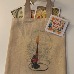 May include: Beige canvas tote bag with a whimsical illustration of a candle and the text "A Dream of Toasted Cheese." The bag contains vintage-style cards and a square sticker that says "Book Tote" with floral accents.
