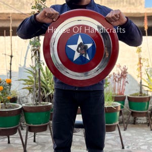 May include: A person holding a red, white, and blue Captain America shield with a silver rim and a five-pointed star in the center. The shield is made of metal and has a textured surface. The text "House Of Handicrafts" is visible on the shield.