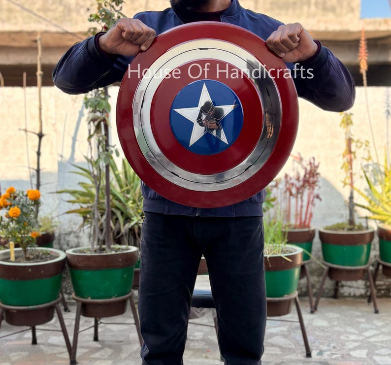 May include: A person holding a red, white, and blue Captain America shield with a silver rim and a five-pointed star in the center. The shield is made of metal and has a textured surface. The text "House Of Handicrafts" is visible on the shield.