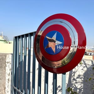May include: A red, white, and blue Captain America shield with a silver metal center and a five-pointed star. The shield is hanging on a metal gate. The text "House Of Handicrafts" is visible in the lower right corner of the image.