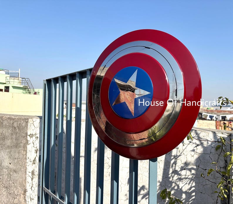 May include: A red, white, and blue Captain America shield with a silver metal center and a five-pointed star. The shield is hanging on a metal gate. The text "House Of Handicrafts" is visible in the lower right corner of the image.