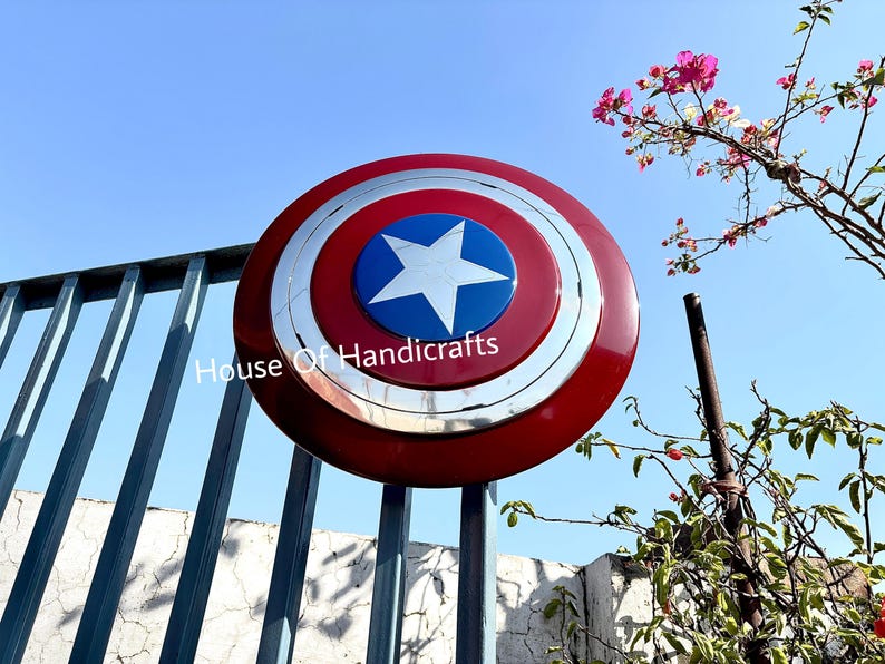 May include: A red, white, and blue Captain America shield with a silver rim. The shield is made of metal and has a star in the center. The shield is hanging on a metal fence.