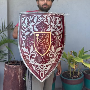 May include: A large, shield-shaped piece with a deep red finish, featuring silver-colored filigree patterns of leaves and vines. A central gold-bordered shield displays a lion holding a hammer. The edge is reinforced with a silver metal trim.