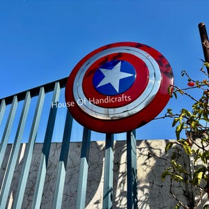 May include: A red, white, and blue Captain America shield with a silver star in the center. The shield is hanging on a metal fence with the text "House Of Handicrafts" visible below it.