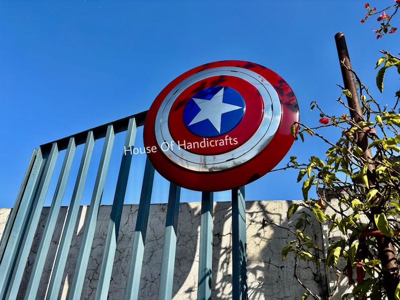 May include: A red, white, and blue Captain America shield with a silver star in the center. The shield is hanging on a metal fence with the text "House Of Handicrafts" visible below it.
