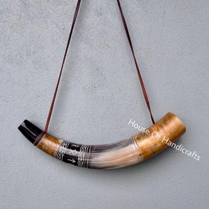 Puede incluir: Un cuerno para beber curvado con un acabado pulido en tonos bronceado y crema, una boquilla negra y una banda negra grabada con símbolos rúnicos blancos y patrones trenzados. Incluye una fina correa de cuero marrón para colgar el cuerno.