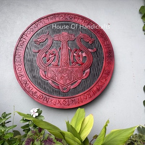 May include: A circular wooden wall plaque featuring a red-stained border with carved runic inscriptions. The centre displays a black-stained background with a relief carving of a hammer flanked by two serpents, with the text House Of Handicrafts.