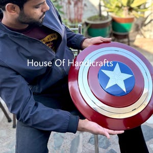 May include: A red, white, and blue Captain America shield with a silver metal rim. The shield has a large white star in the center. The shield is being held by a person. House Of Handicrafts.