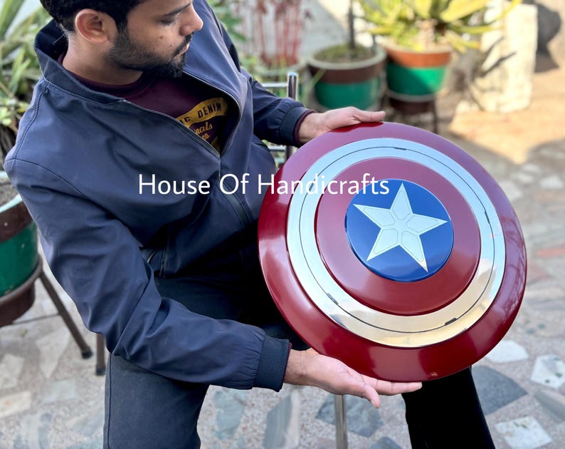 May include: A red, white, and blue Captain America shield with a silver metal rim. The shield has a large white star in the center. The shield is being held by a person. House Of Handicrafts.